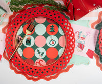 Overhead place setting featuring a red scalloped paper charger, a retro green and red dot pattern plate, and a gold fork.