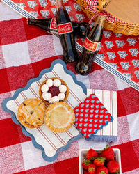 Red block print cocktail napkin and blue scalloped dinner plate with mini pies on red gingham picnic blanket