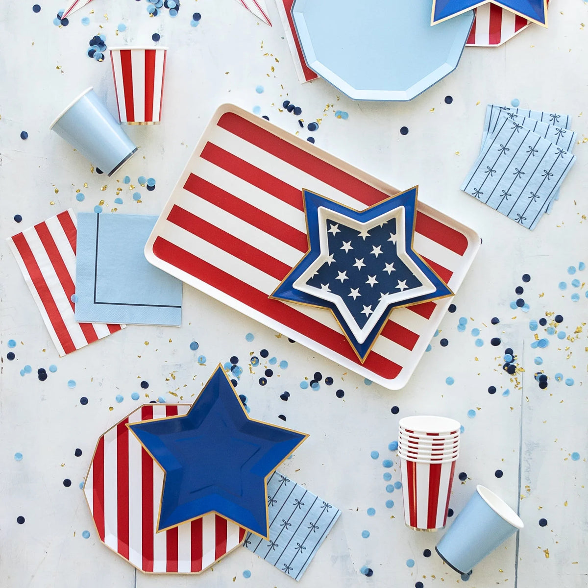 Fourth of July party supplies flat lay with red and white striped flag tray, navy star shaped plates, striped paper cups, and red striped napkins with blue confetti.