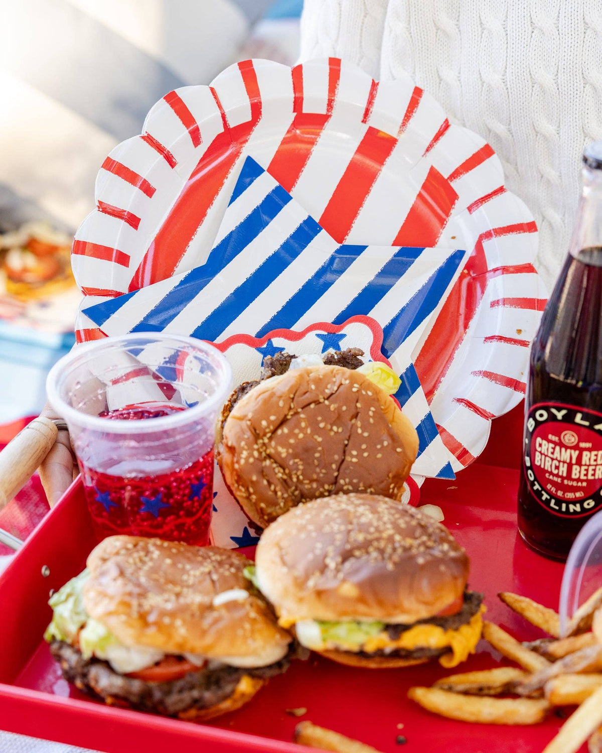 Red striped scalloped paper dinner plate with blue striped napkin on a red serving tray with burgers, fries, and a patriotic cup