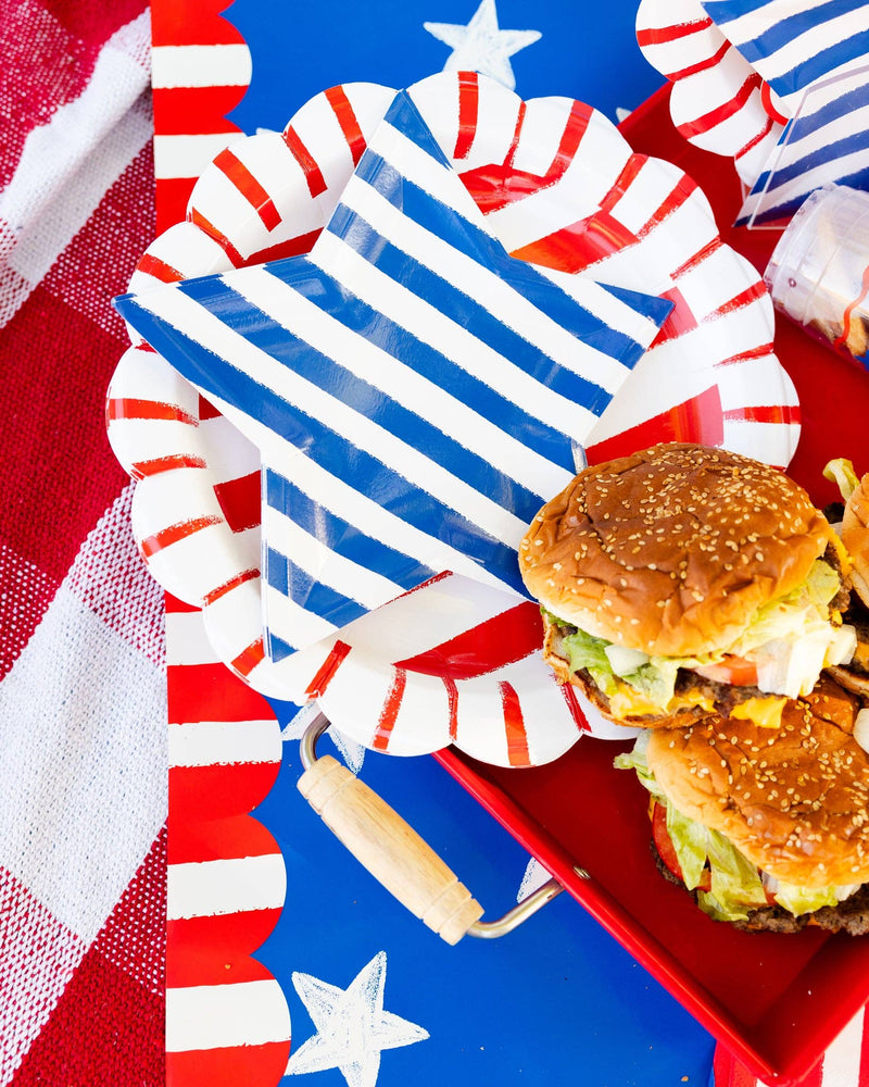 Overhead view of red striped and blue striped scalloped paper plates layered on a patriotic table runner with burgers on a red tray