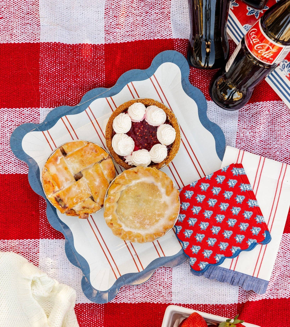 Red striped scalloped paper plate with mini pies and red block-print floral napkin on red and white gingham picnic blanket — summer Americana styling