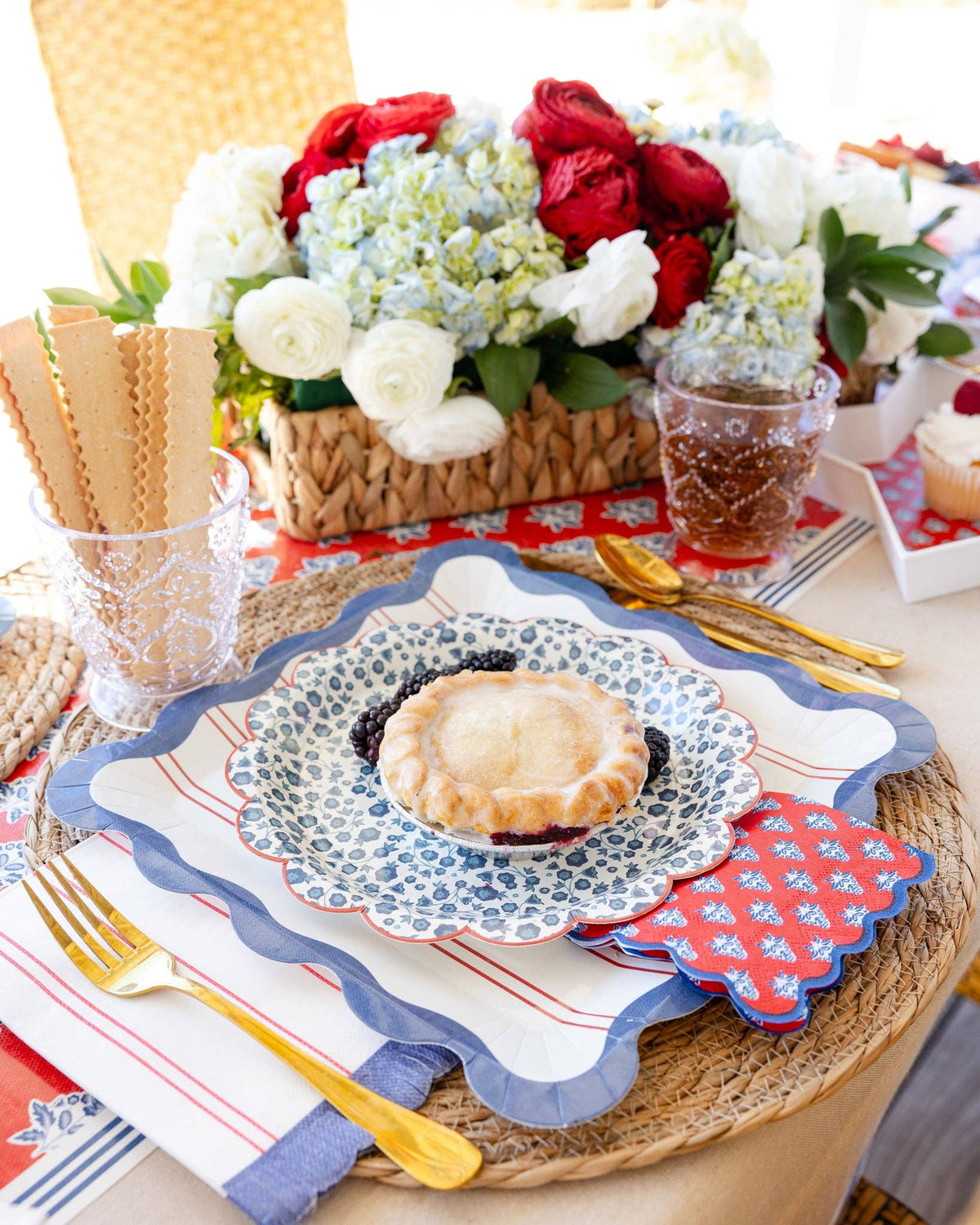 Red striped scalloped paper dinner plate layered with blue floral salad plate, mini pie, and red block-print napkin on wicker placemat with gold cutlery and Hamptons Americana table setting
