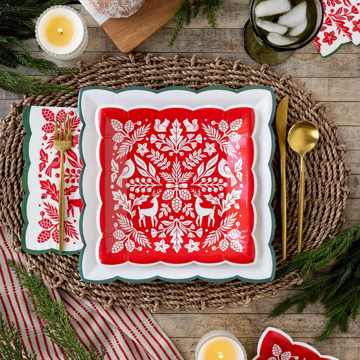 Red woodland paper dinner napkin with a green scalloped edge beside a red folk art square plate on a woven placemat with gold flatware
