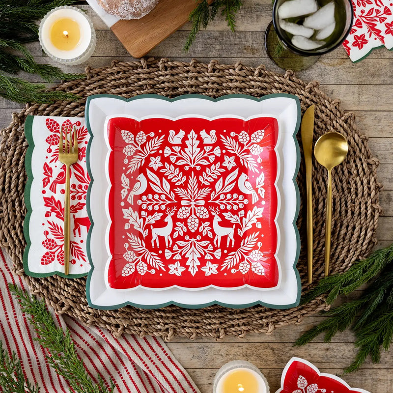 Red woodland paper dinner napkin with a green scalloped edge beside a red folk art square plate on a woven placemat with gold flatware