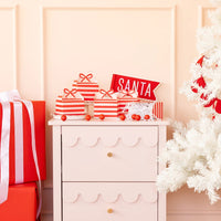 Display of assembled Christmas cookie boxes on a pink cabinet with a white tree, showcasing the red and pink party supplies palette.