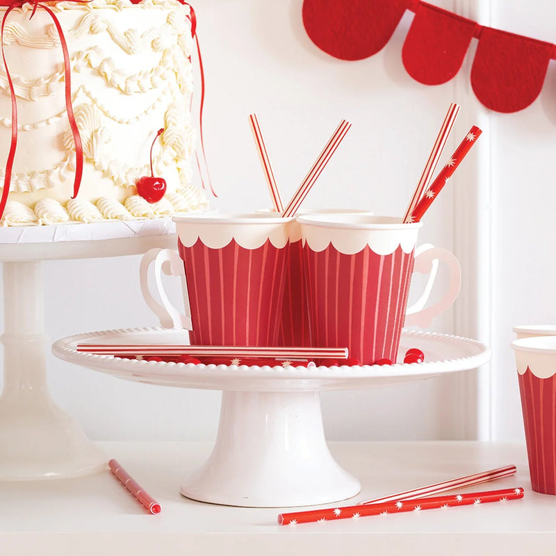 Close-up of red straws placed in striped party cups on a white cake stand, perfect for holiday party supplies.