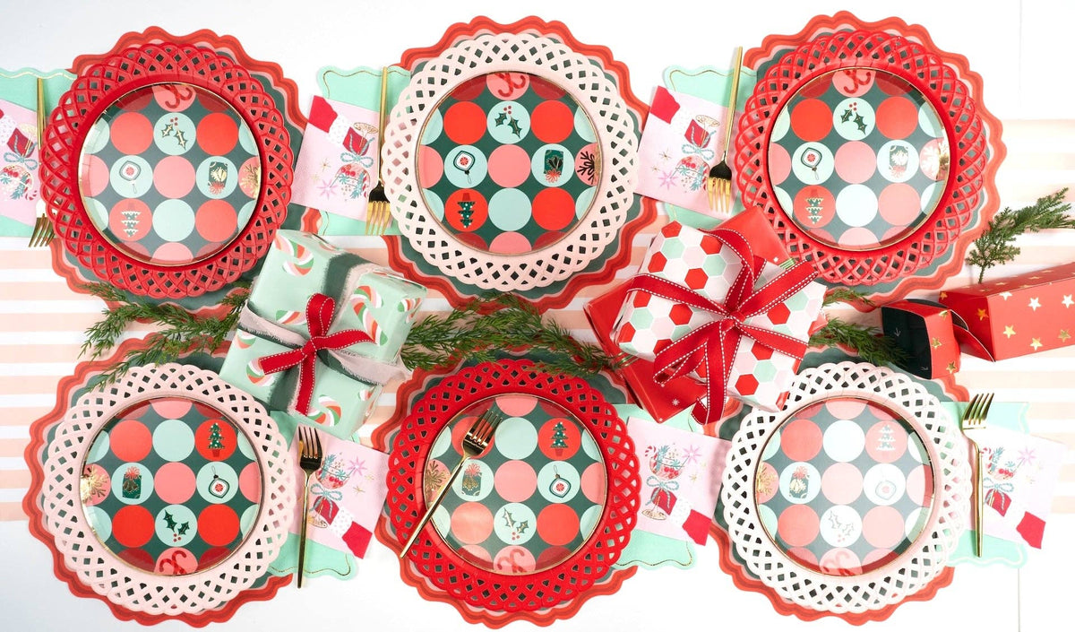 Wide overhead flat lay of a holiday table setting packed with red scalloped chargers, dot plates, pink napkins, and small wrapped presents.