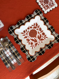 Overhead fall place setting with a white square scalloped paper plate in a rust folk floral print on a black and white plaid placemat, a pink glass, and gold flatware on a rust orange tablecloth