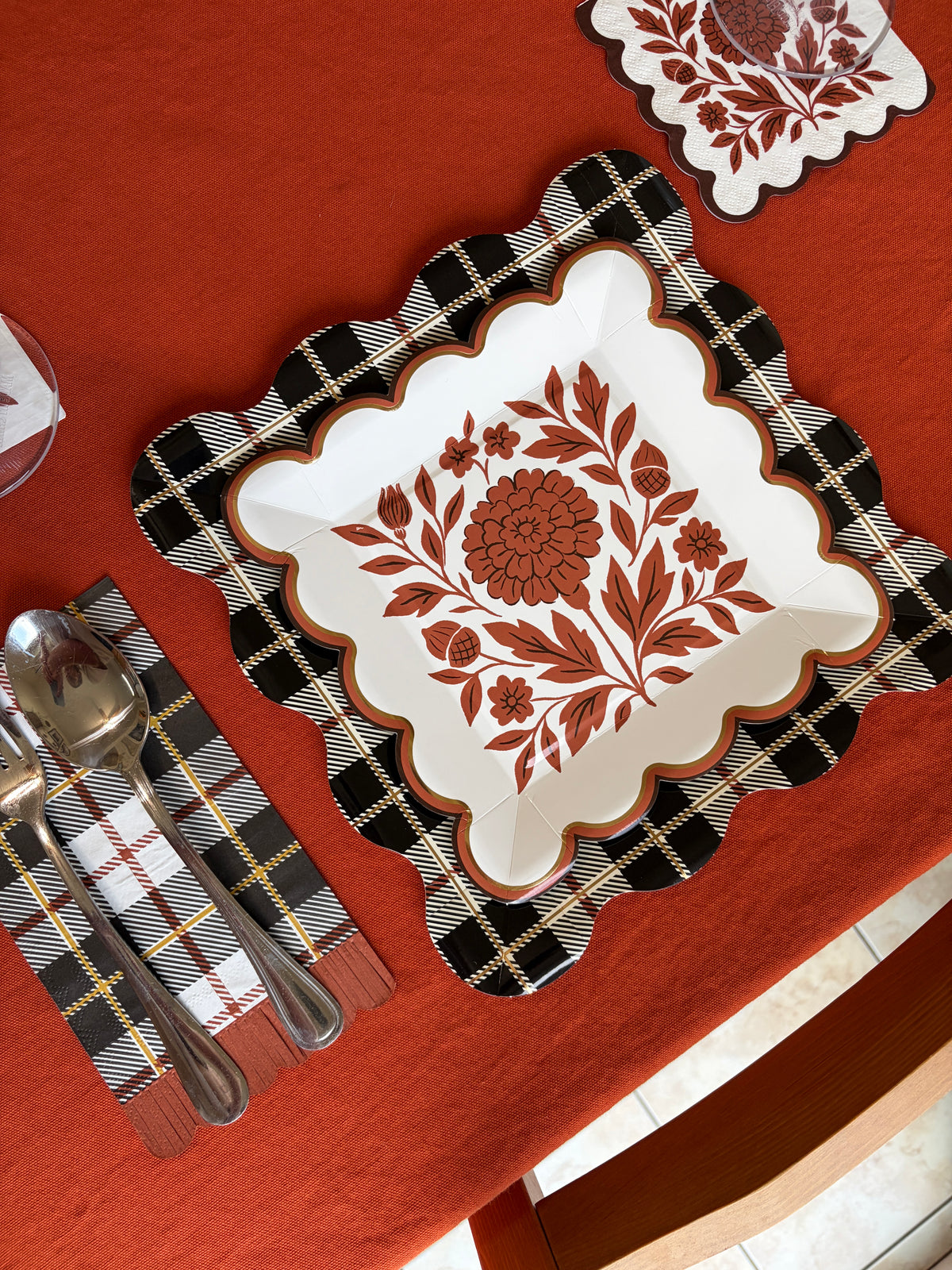 Overhead fall place setting with a white square scalloped paper plate in a rust folk floral print on a black and white plaid placemat, a pink glass, and gold flatware on a rust orange tablecloth