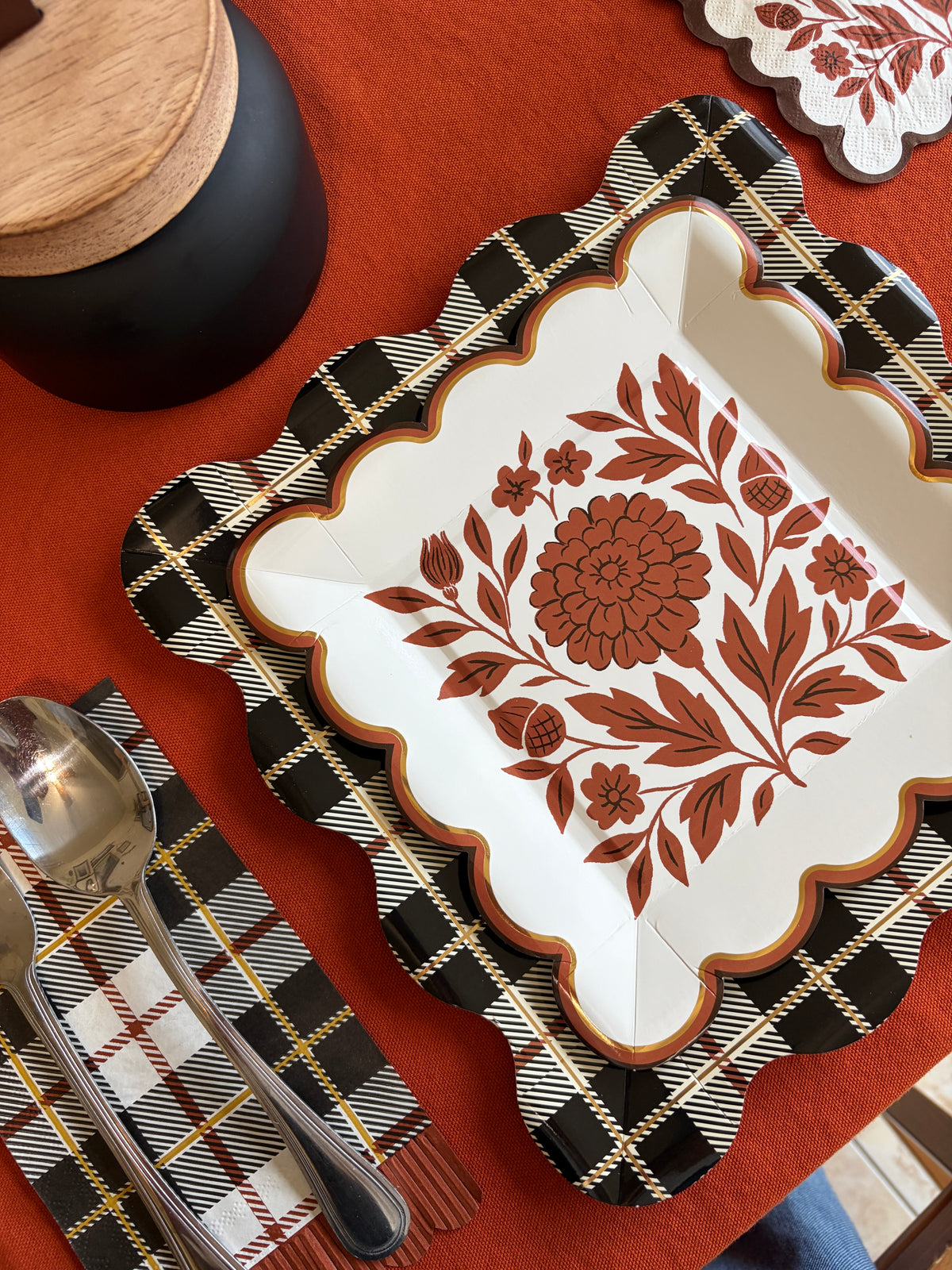 Overhead close-up of a rust folk floral square paper plate on a black and white plaid napkin with gold flatware on an orange tablecloth