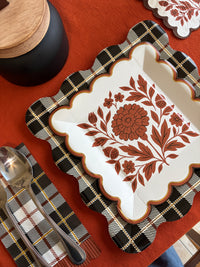 Overhead close-up of a rust folk floral square paper plate on a black and white plaid napkin with gold flatware on an orange tablecloth