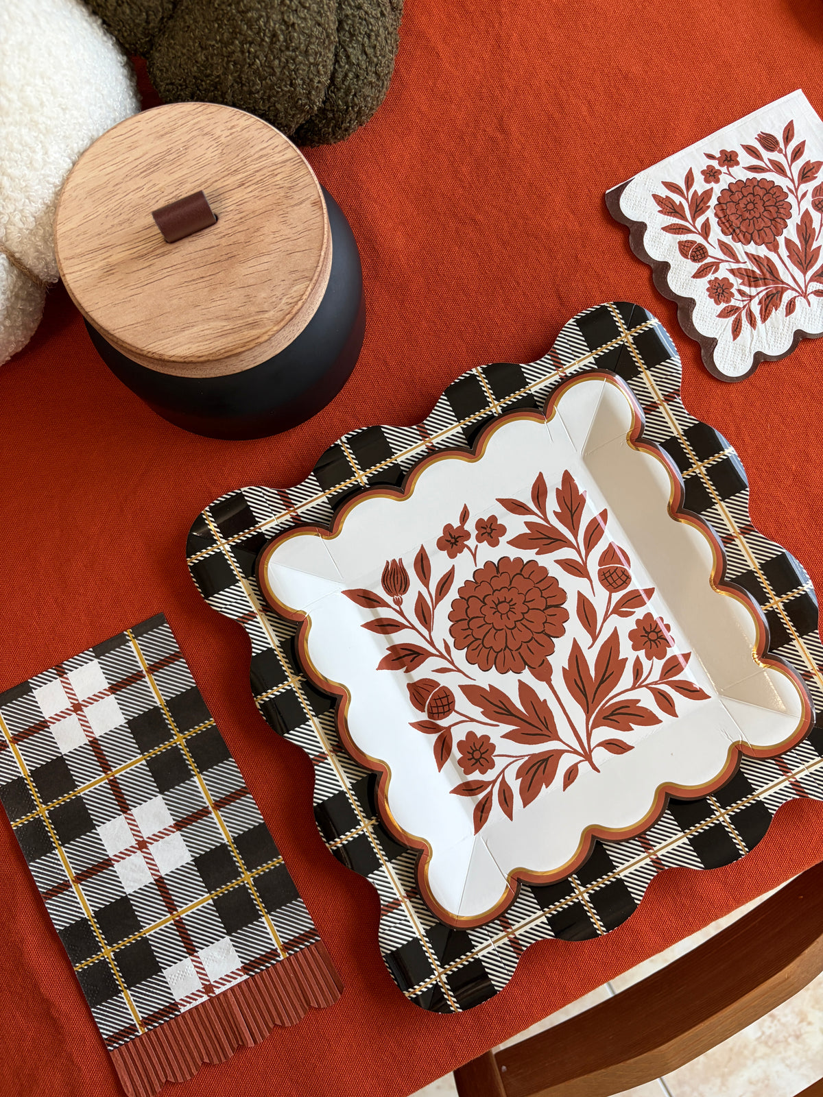 Angled fall place setting with a rust folk floral square paper plate on a black and white plaid placemat, a scalloped napkin, knit pumpkins, and a wood-lidded black candle
