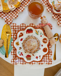 Thanksgiving-themed table setting featuring rust gingham charger plate with other decorative plates, cutlery, and a glass of juice.