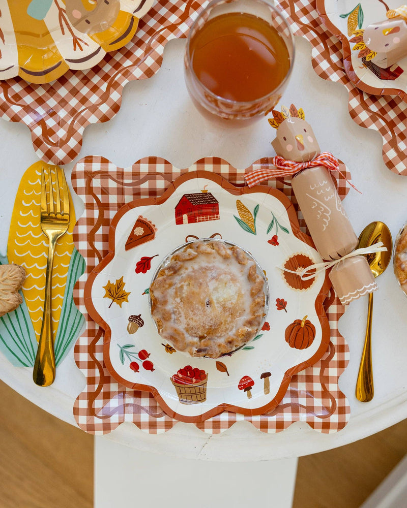 Thanksgiving-themed table setting featuring rust gingham charger plate with other decorative plates, cutlery, and a glass of juice.