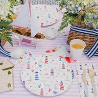 Sandcastle shaped paper plates with gold foil detailing on a pink striped tablecloth beside a seaside print round paper plate, lighthouse napkins, and a toy sailboat at a beach birthday party.