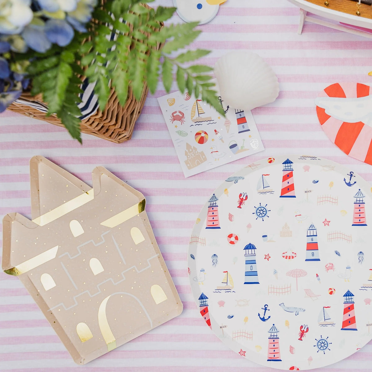 Flat lay of a sandcastle shaped paper plate next to a seaside print paper dinner plate, beach sticker sheet, and striped napkins on a pink and white striped table linen.