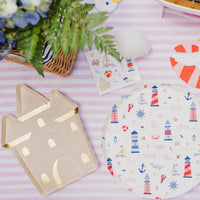 Flat lay of a sandcastle shaped paper plate next to a seaside print paper dinner plate, beach sticker sheet, and striped napkins on a pink and white striped table linen.
