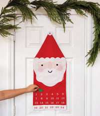 Santa canvas advent calendar hung on a white door with a hand reaching for a numbered pocket