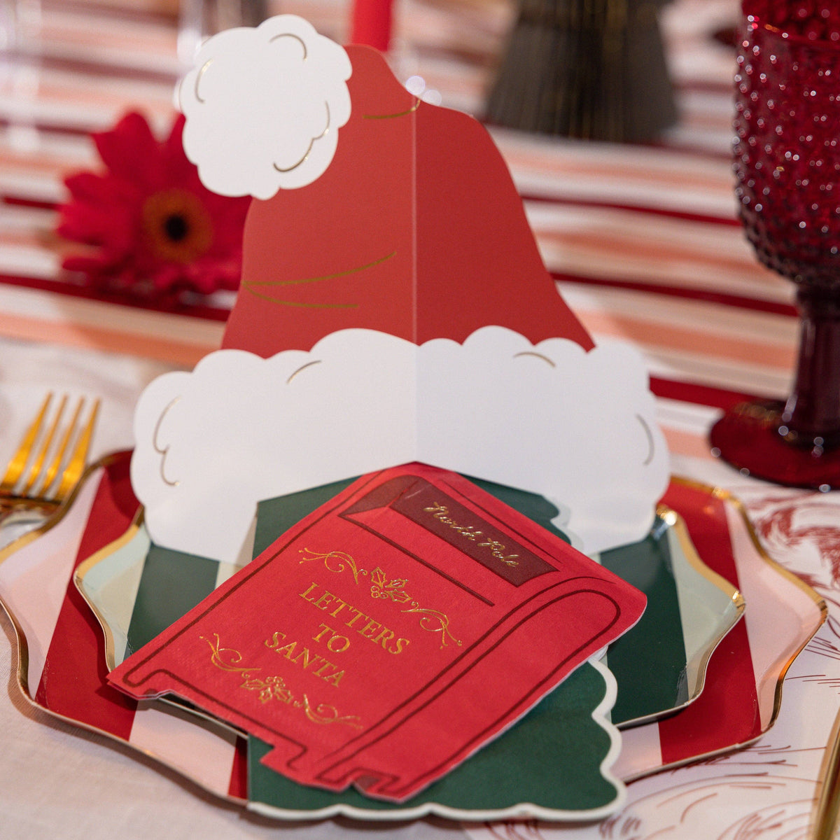 Close-up of a folded red Santa-hat place card with white trim, set on green Christmas-tree plates over a striped runner beside a red goblet.