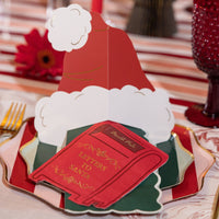 Close-up of a folded red Santa-hat place card with white trim, set on green Christmas-tree plates over a striped runner beside a red goblet.