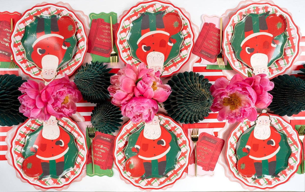 Overhead of Santa plates and red gift-card napkins on plaid plates around pink peonies and fringe trees.