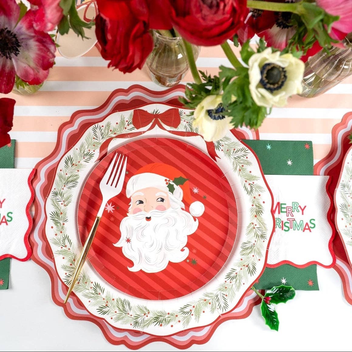 Place setting with the Santa dessert plate on a wreath plate and charger, a Merry Christmas banner, striped runner, and red flowers.