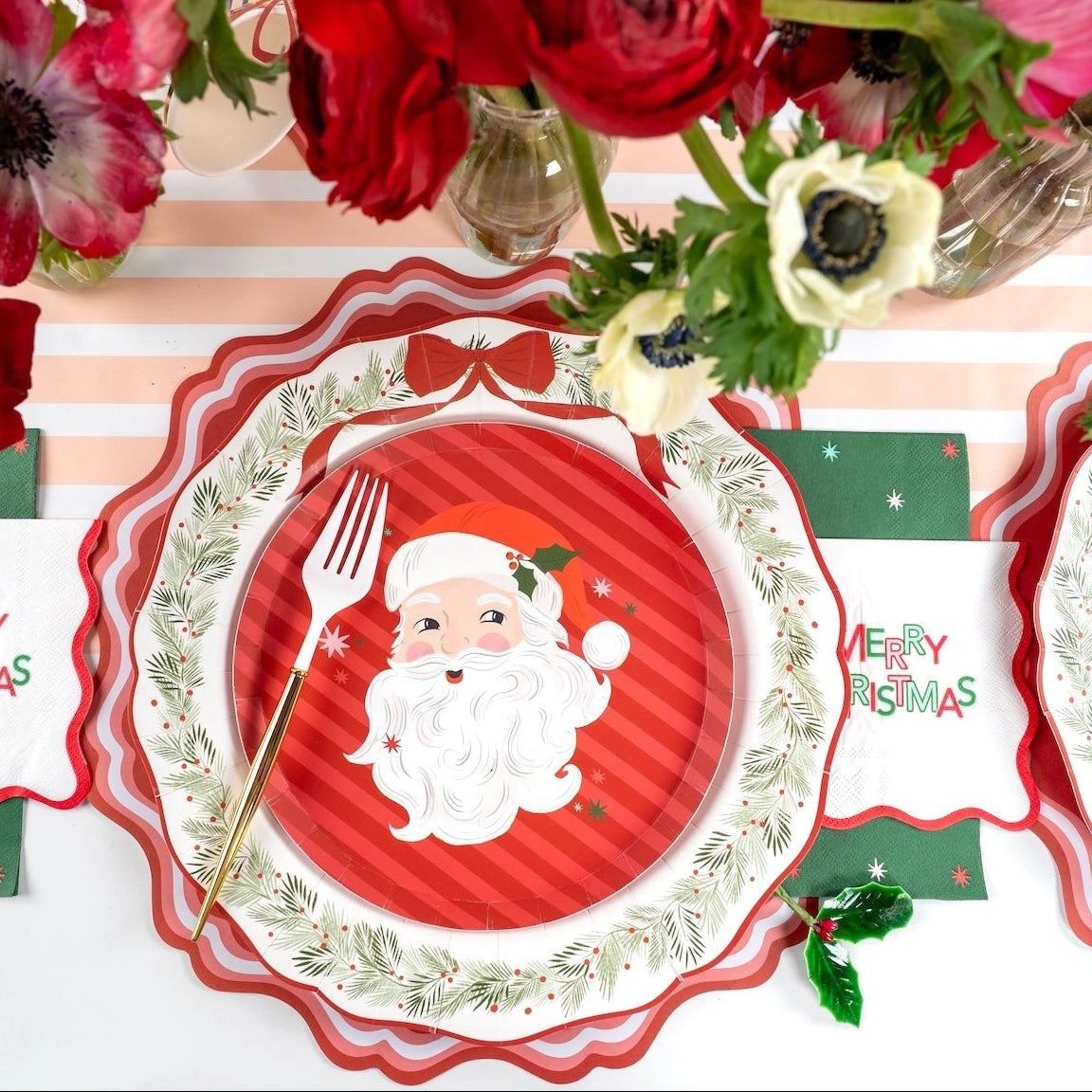 Place setting with a Santa plate on a scalloped plate, the green star guest towel, red flowers, and a Merry Christmas banner.