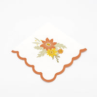 Angled view of a white scalloped cocktail napkin with a rust border and fall flower print