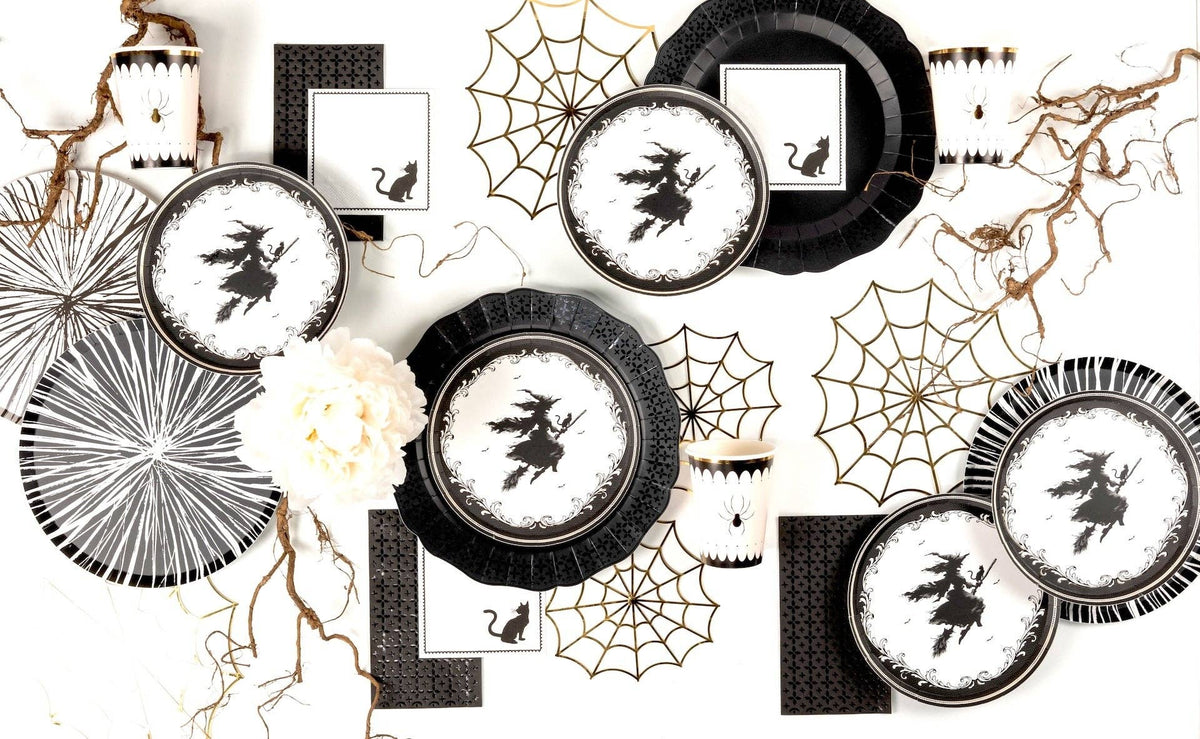 Scattered tablescape flat lay displaying multiple vintage witch paper plates, black scalloped chargers, and bold paper spiderwebs.