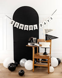 School Days felt banner hung across a black arch backdrop with black and white balloons, wooden school chair, white layer cake, and 1st Day of School and Last Day of School pennants