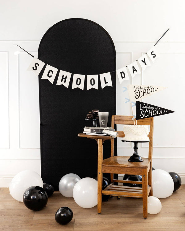 School Days felt banner hung across a black arch backdrop with black and white balloons, wooden school chair, white layer cake, and 1st Day of School and Last Day of School pennants