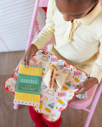 Young girl in yellow polo holding school supplies scalloped paper plate with PB&J sandwich and pencil box paper napkin