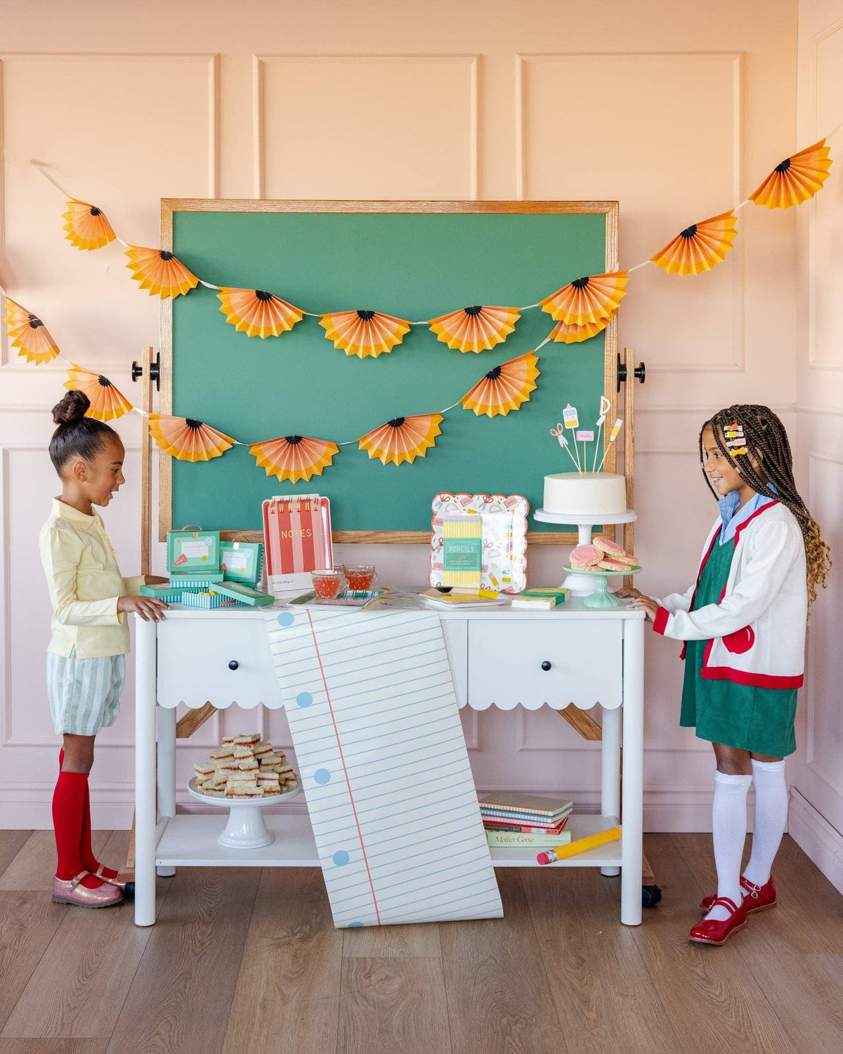 Styled back to school party with two children at white desk with notebook paper runner, pencil shaving banner, chalkboard backdrop, cake, and school supplies scalloped plate