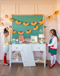 Styled back to school party with two children at white desk with notebook paper runner, pencil shaving banner, chalkboard backdrop, cake, and school supplies scalloped plate