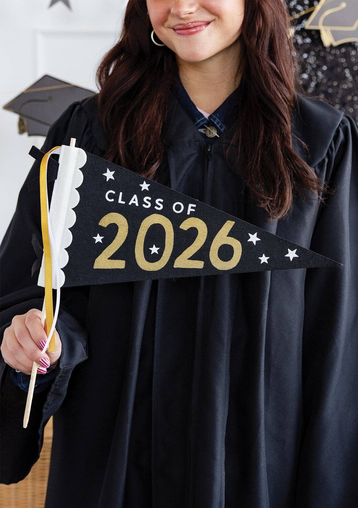 A high school graduate in a black cap and gown holding a Class of 2026 Sign as a photo backdrop prop.