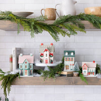 Wide shot of a shelf displaying the full lighted Christmas village collection, decorated with greenery and faux snow.