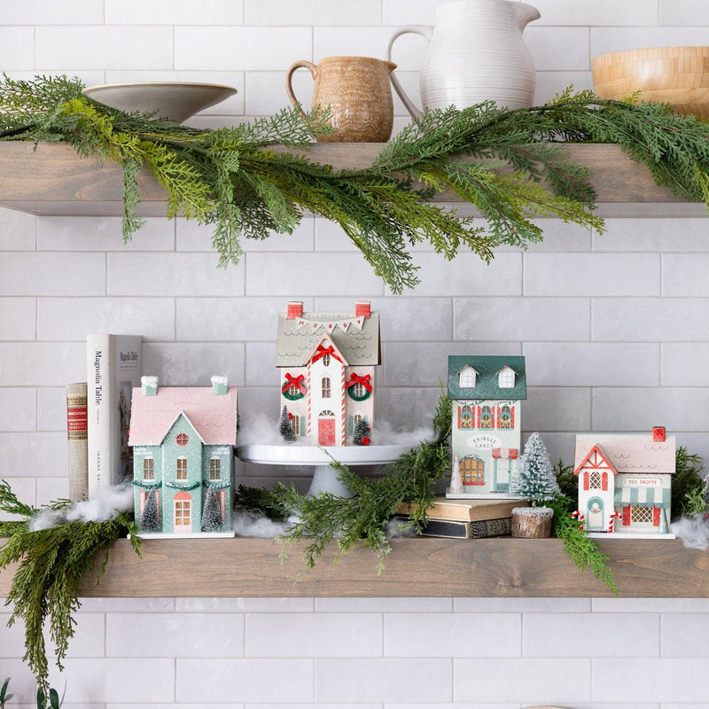 Wide shot of a shelf displaying the full lighted Christmas village collection, decorated with greenery and faux snow.