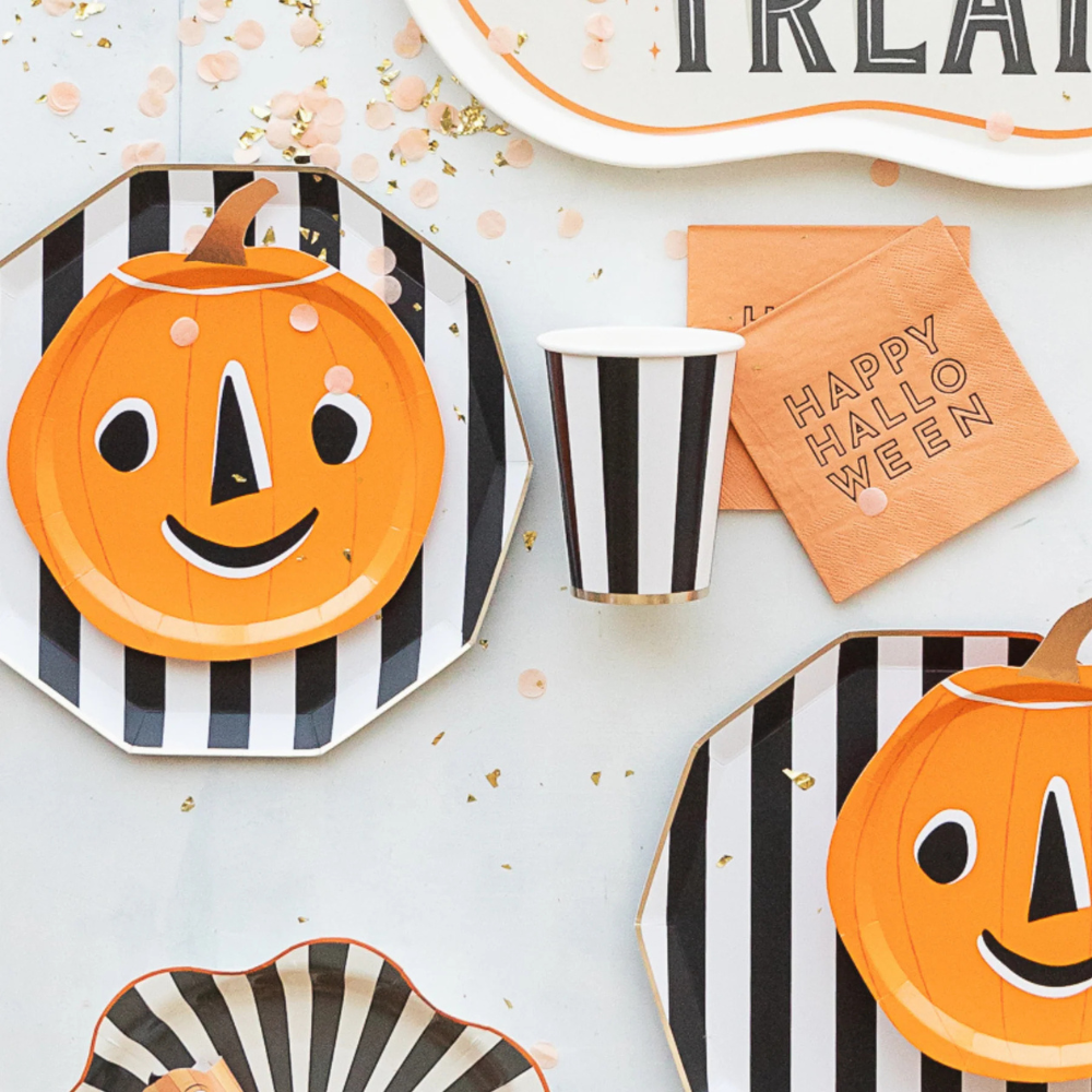 Halloween tableware flat lay with black-and-white striped plates, pumpkin plates, striped cups, and orange cocktail napkins.
