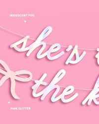 She's Tying the Knot banner showing iridescent foil lettering and pink glitter bow material details on pink background