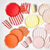 Party supplies flat lay with red, orange, yellow, blush, and pink striped and solid paper plates, striped paper cups, and starburst print napkins on a white surface.