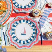 Sky blue striped-bow dinner plate layered with a navy stripe sailboat plate, beach club napkin, and blue cutlery on a red stripe runner.