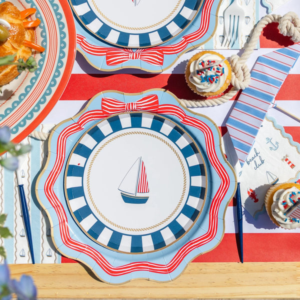 Sky blue striped-bow dinner plate layered with a navy stripe sailboat plate, beach club napkin, and blue cutlery on a red stripe runner.