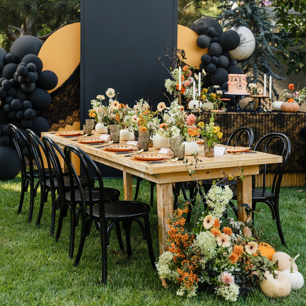 Outdoor Halloween dinner party table with orange spiderweb place settings, fall florals, white pumpkins, and black balloon garlands