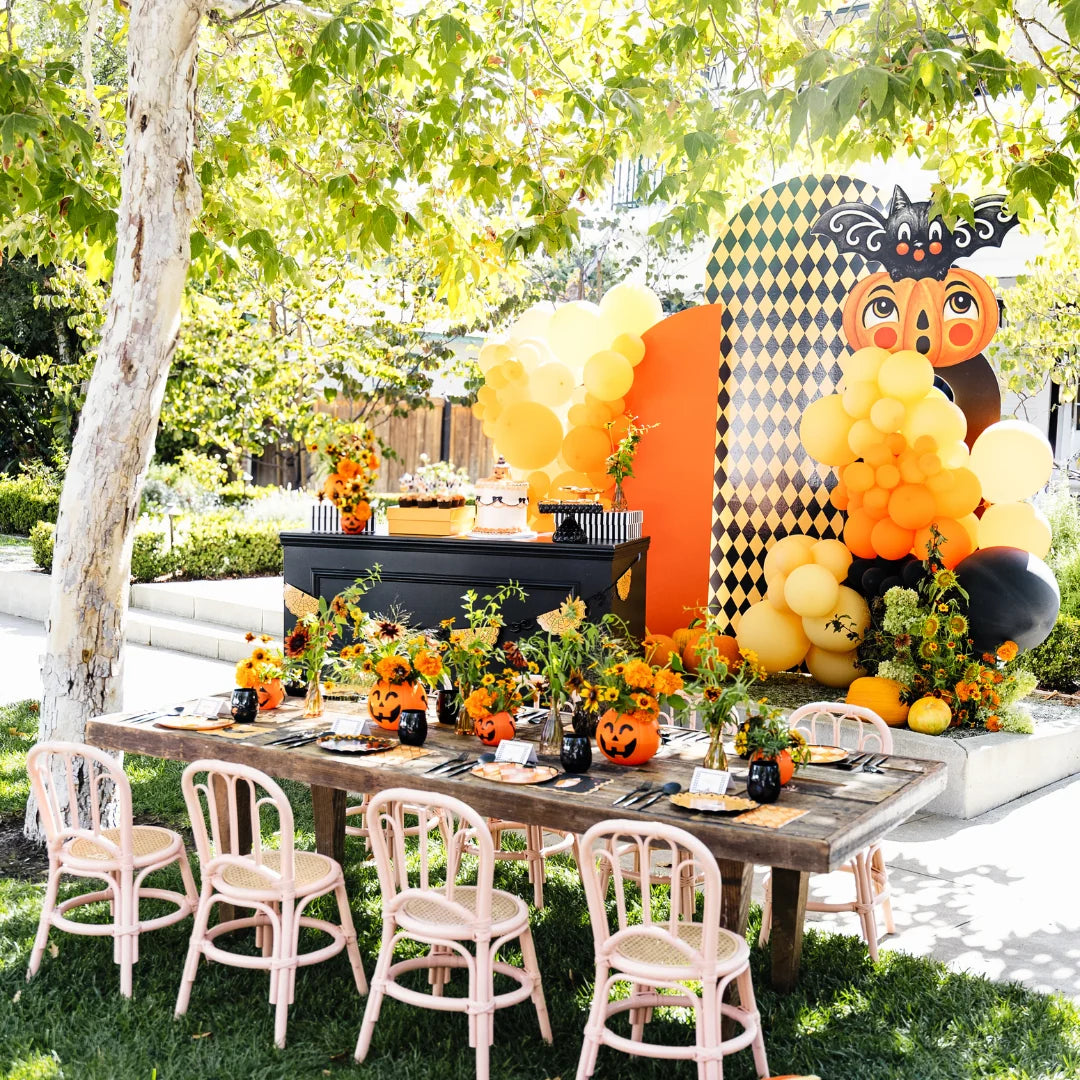 Vintage Halloween kids party table set outdoors with jack-o'-lantern centerpieces, pink rattan chairs, and an orange and black balloon backdrop