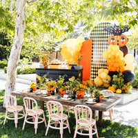 Vintage Halloween kids party table set outdoors with jack-o'-lantern centerpieces, pink rattan chairs, and an orange and black balloon backdrop
