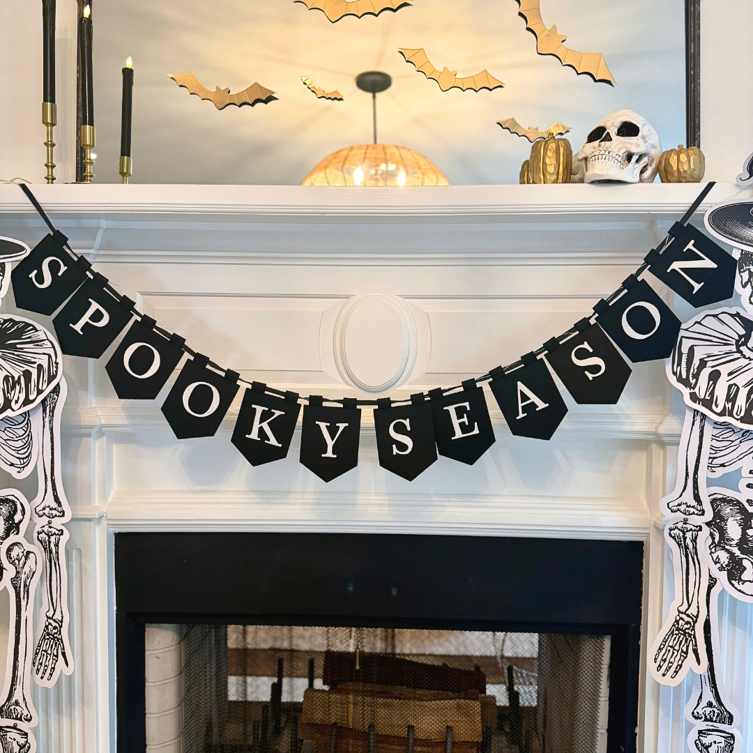 Close-up of a black "Spooky Season" pennant banner with white letters draped across a white mantel, with gold paper bats, a skull, and gold pumpkins on the mantel above.