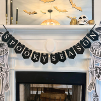 Close-up of a black "Spooky Season" pennant banner with white letters draped across a white mantel, with gold paper bats, a skull, and gold pumpkins on the mantel above.
