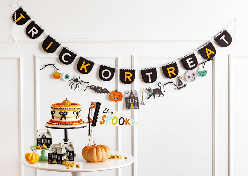 Straight-on shot of a white paneled wall above a round white dessert table. The character paper banner hangs below the Trick or Treat felt banner. All 11 die-cut characters are visible across the garland span: candy corn, eyeball, spider, bat, jack-o-lantern, haunted house, broom, black cat, dancing figure, skull, and potion bottle. Black velvet bows visible at intervals. 