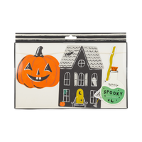 Front of retail packaging showing three visible die-cut pieces inside a windowed box: a large orange jack-o-lantern with a friendly carved face, a black haunted house with gray scalloped roof and yellow-lit doorway (ghost visible in an upper window, spider on rooftop), and a green potion bottle with a broom handle protruding from the top. Small pumpkin sticker label reads "SPOOKY Paper Banner."